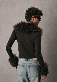 Maglione marrone a coste con colletto e polsini in pelliccia soffice, abbinato a jeans chiari strappati. I dettagli testurizzati mettono in risalto il design.