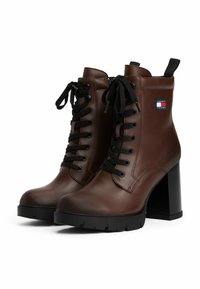 Bottes chelsea en cuir marron avec des lacets noirs, bout rond, talon épais noir, et logo Tommy Jeans sur le côté. Détail de surface texturé.