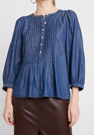 Mujer vistiendo una blusa azul de manga larga con frente plisado y botones blancos, combinada con una falda de cuero marrón.