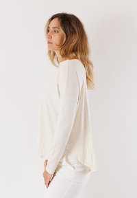 Maglietta a maniche lunghe color crema chiaro con vestibilità rilassata, orlo arrotondato e texture morbida. Il materiale sembra leggero e traspirante.