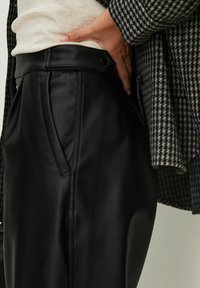 Hand in der Tasche von schwarzen Lederhosen, kombiniert mit einem beigen Oberteil und einer schwarz-weißen karierten Jacke.