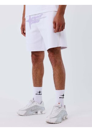 Hombre vistiendo pantalones cortos blancos, calcetines blancos hasta la mitad de la pantorrilla con logo negro, y zapatillas deportivas blancas con detalles grises, de pie sobre un suelo blanco.