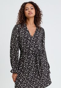 Robe noire portefeuille avec motif floral blanc, décolleté en V profond, manches longues, taille cintrée avec lien à nouer et poignets volants. Tissu léger avec une texture douce.