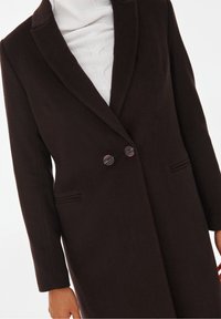 Manteau en mélange de laine marron avec un col cranté, fermeture à double bouton à l'avant, deux poches latérales et des détails brodés le long des poches.