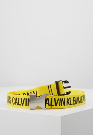 Gelber Stoffgürtel mit schwarzem "CALVIN KLEIN JEANS"-Schriftzug und silberner Metallschnalle, auf weißer Oberfläche vor einfarbigem Hintergrund gelegt.