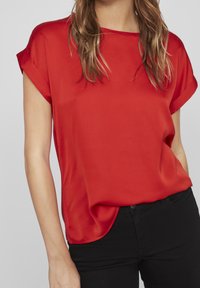 Blusa rossa a maniche corte con una vestibilità ampia, texture liscia e scollatura rotonda. Abbinata a pantaloni neri aderenti, che enfatizzano la semplicità.