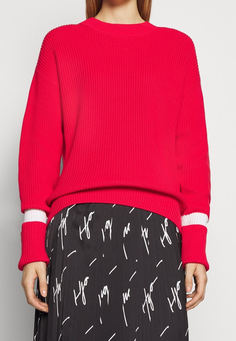 Un pull rose vif côtelé à manches longues avec des poignets rayés blancs, associé à une jupe noire ornée d'un motif textuel blanc.