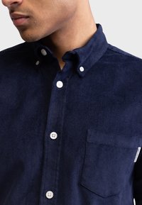 Camisa de pana de color marino con cuello abotonado, bolsillo delantero y botones blancos. La tela tiene una textura suave y acanalada verticalmente.