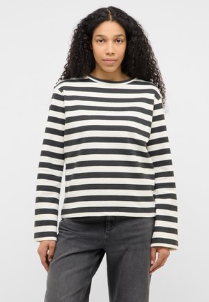 Femeie cu păr lung și creț, purtând o bluză cu mânecă lungă cu dungi orizontale alb-negru și blugi gri închis, stând în picioare pe un fundal simplu.