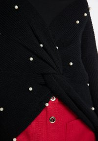 Cardigan nero lavorato a maglia con design intrecciato e decorazioni di perle, indossato sopra una camicia rossa texturizzata con bottoni decorativi.