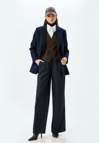 Marineblauw dubbelbreasted jasje over een bruine vest, gecombineerd met een wijd-uitlopende donkere broek. Een bruine pet en puntige zwarte hakken maken de look compleet.