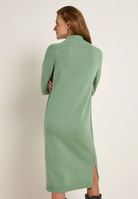 Grünes Strickkleid mit hohem Kragen, langen Ärmeln und seitlichen Schlitzen. Rippendetails am Saum und Kragen. Weicher Stoff.