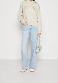 Přesměrně velká béžová mikina s kovovým logem "GAP", světle modré široké džíny a malá kabelka mátové barvy. Bílé tenisky s hnědými podrážkami.