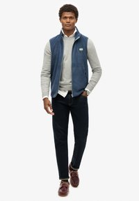 Granatowy polarowy kamizelka nałożona na jasnoszary, długi rękaw, w zestawieniu z ciemnymi dżinsami i brązowym obuwiem. Zawiera logo na piersi oraz boczne kieszenie.