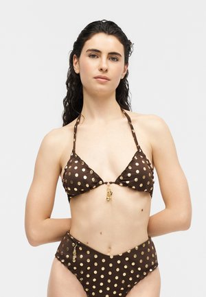 Donna che indossa un bikini marrone a halter con pois dorati e piccoli dettagli con ciondolo dorato sulla parte superiore e inferiore, su uno sfondo chiaro e semplice.