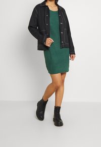 Veste noire à boutons avec des coutures blanches sur une robe en maille côtelée verte. Mannequin portant des bottes chelsea noires à lacets.