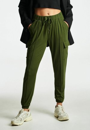 Personne portant un pantalon cargo jogging vert olive avec taille à cordon, un top court noir et des baskets blanches, debout les mains dans les poches.