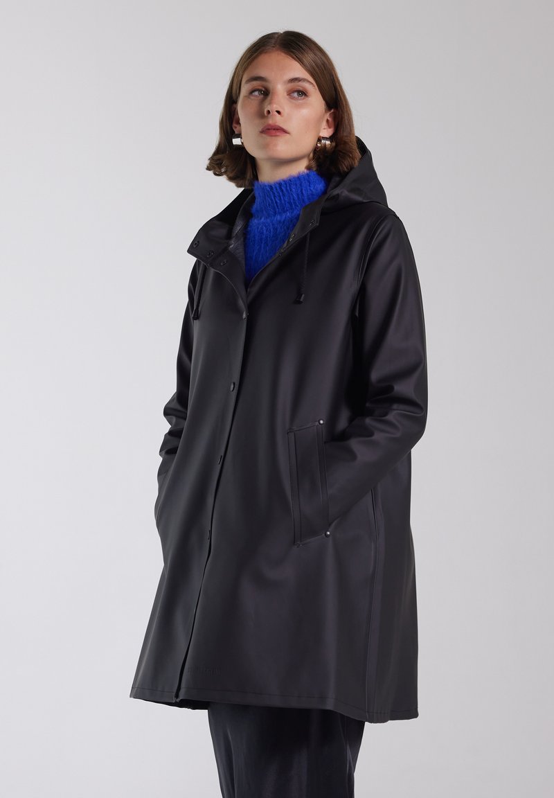 Svart vattentät regnjacka med huva, tryckknappar och sidofickor. Bärs över en blå texturerad tröja. Slät ytfinish.