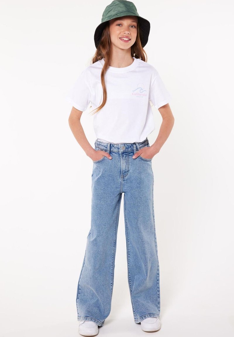 Jeune fille portant un t-shirt blanc à motif, un jean bleu ample, des baskets blanches et un chapeau bob vert, debout les mains dans les poches.