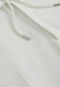 Fehér anyag, sima textúrával és zsinóros részletezéssel. A zsinór ezüst fém véggel rendelkezik, ami minimalista design elemet ad hozzá.