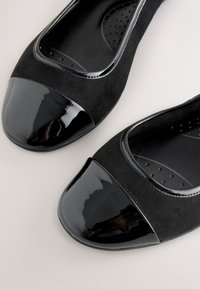 Paar schwarze flache Schuhe mit glänzenden Lackkappen und weichem Wildlederkörper, von oben auf hellem Hintergrund gezeigt.
