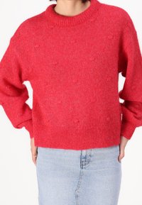 Jasnoczerwona dzianinowa sweter z zaokrąglonym dekoltem, opadającymi ramionami i teksturowanymi detalami w formie pomponów; w zestawie z jasną spódnicą jeansową.