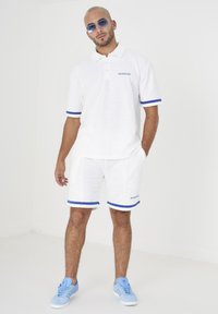 Polo shirt bianco testurizzato e pantaloni corti abbinati con accenti e ricami blu. Abbinato a sneakers azzurre e occhiali da sole.