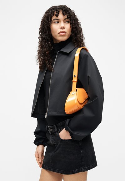 Veste courte noire avec fermeture éclair, associée à une mini-jupe noire. Tient un sac à main orange brillant au design structuré avec une bandoulière.