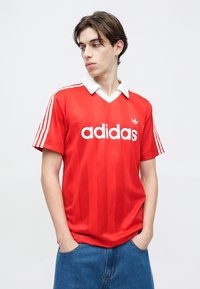 Νέος άνδρας με τα χέρια στις τσέπες, φοράει κόκκινο πουκάμισο Adidas με λευκό γιακά και ρίγες, κοιτάζοντας προς τα πλάγια μπροστά σε απλό φόντο.