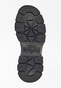 Black rubber shoe sole with a textured surface, featuring a geometric pattern, grooves, and a triangular logo marked "GUESS."