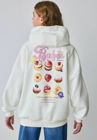 Valge ülemsuuruses kapuutsiga sviiter, millel on seljal värviline magustoidu graafika, sealhulgas kooke ja saia, ning tekst "Baker by Ted Baker".
