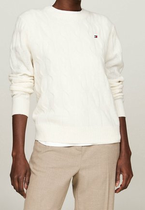 Persona vistiendo un suéter blanco de punto trenzado con mangas remangadas y pantalones beige, mostrada desde el cuello hasta las caderas sobre un fondo neutro.