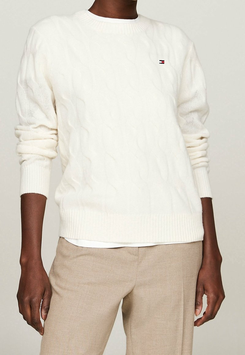 Personne portant un pull blanc en maille torsadée avec les manches retroussées et un pantalon beige, montrée du cou jusqu'aux hanches sur un fond neutre.
