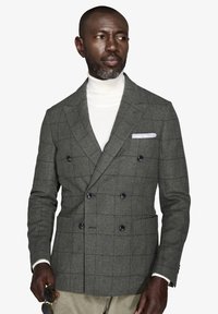 Gråternet dobbeltradet jakke i struktureret uld, med sorte knapper, en hvid turtle-neck trøje og et dekorativt lommetørklæde.