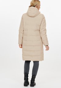 Beige puuvillane mantel, millel on kapuuts, sile tekstuur, pikkus ja nupud. Seljas hallid teksad ja mustad saapad.