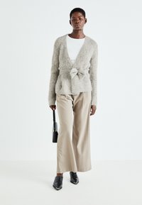 Cardigan fuzzy taupe cu un nod la talie, asortat cu pantaloni largi bej deschis și mule negre. Geantă minimalistă neagră în mână.