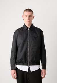 Veste à carreaux noirs avec fermeture éclair à l'avant, col à pression, taille élastique et une texture sur le patch à gauche de la poitrine; portée sur une chemise blanche.