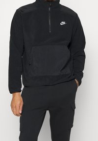 Schwarzer Nike-Pullover mit halbem Reißverschluss-Kragen, strukturierter Stoff, Fronttasche und Logo auf der Brust. Kombination mit passenden schwarzen Hosen.