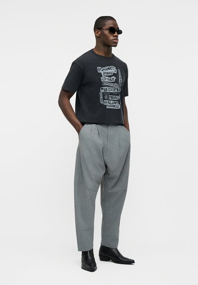 Άνδρας με μαύρο μπλουζάκι με σχέδιο, γκρι πλισέ παντελόνι, μαύρες μπότες και γυαλιά ηλίου, στέκεται με τα χέρια στις τσέπες μπροστά σε λευκό φόντο.