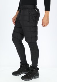 Zwarte geïsoleerde shorts met een gewatteerd ontwerp, voorzien van zijritszakken, gedragen over donkergrijze leggings en zwarte wandelschoenen.