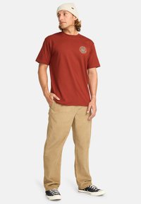 T-shirt en coton rouge avec un logo circulaire sur la poitrine, associé à un pantalon en velours côtelé khaki et des baskets noires avec des accents blancs.