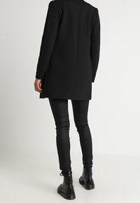 Manteau long noir texturé à coupe droite, accompagné d'un pantalon skinny noir et de bottines à la cheville. Design simple sans motifs visibles.