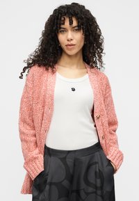 Cardigan tricotat într-o culoare coral texturată, cu mâneci lungi și nasturi aurii. Purtat peste un top alb cu un mic desen floral negru.