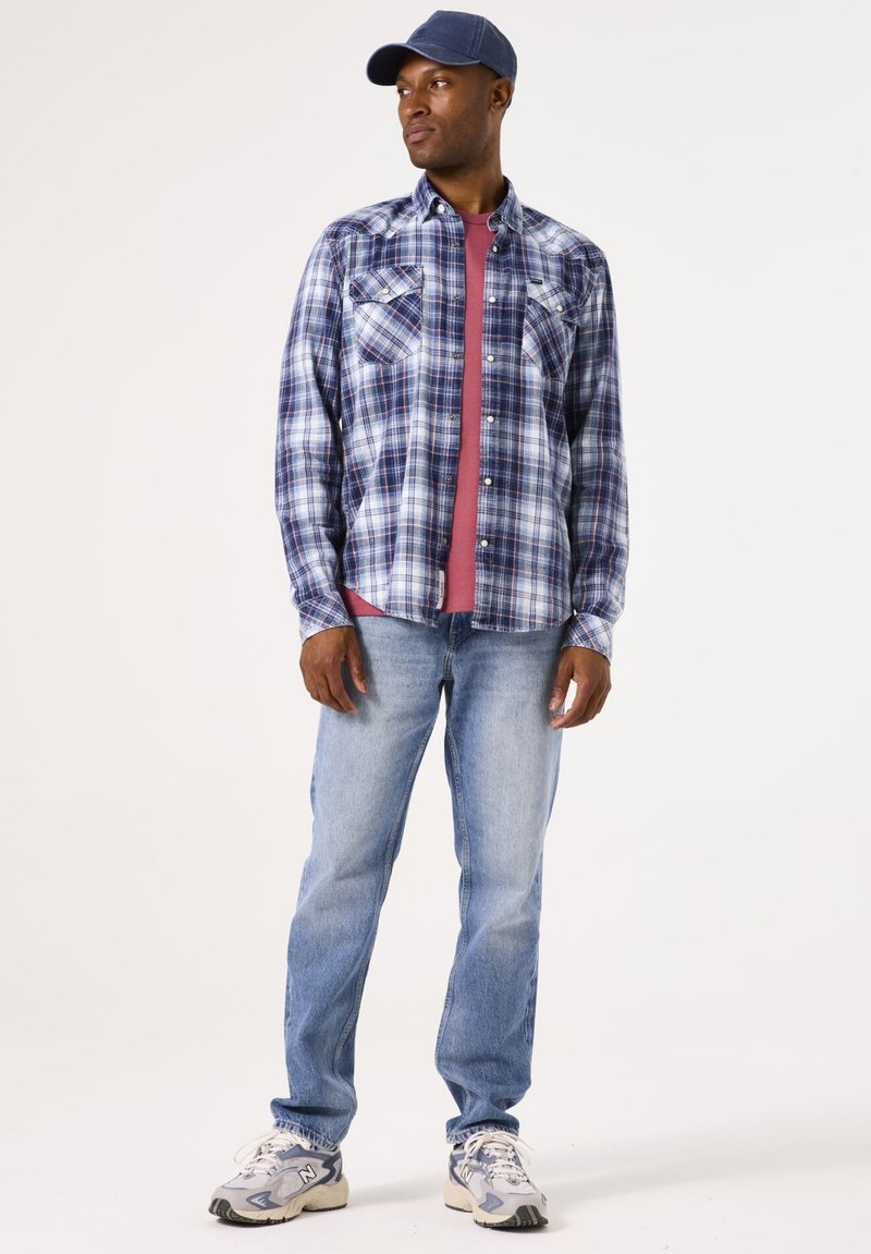 Camisa de cuadros azul, blanca y roja, superpuesta a una camiseta color marrón. Combinada con jeans de un azul claro y zapatillas deportivas grises.