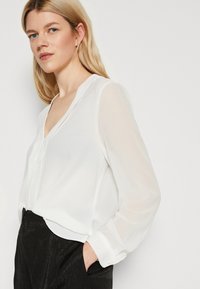 Blusa bianca a maniche lunghe con scollatura a V e dettagli con bottoni, realizzata in un materiale traslucido, abbinata a pantaloni neri con texture.