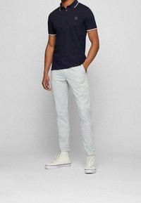 Polo shirt blu navy con accenti bianchi, maniche corte e taschino quadrato con logo, abbinato a pantaloni slim-fit grigio chiaro e sneaker alte bianche.