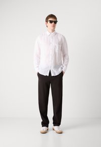 Camisa blanca de lino con botones y bolsillo, pantalones negros holgados y zapatillas blancas con acentos beige. Modelo llevando gafas de sol oscuras.