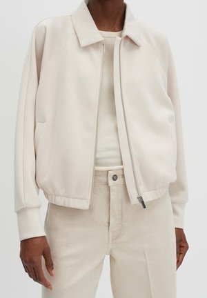 Personne portant une veste beige clair zippée sur un haut côtelé et un pantalon beige, montrée des épaules aux hanches sur un fond uni.