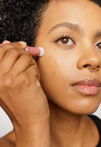 Applikatorstift in der Hand, trägt das Produkt mit einer weißen Applikatorspitze auf die Wange auf. Die Haut wirkt glatt, mit einem natürlichen Glow und minimalen Unreinheiten.