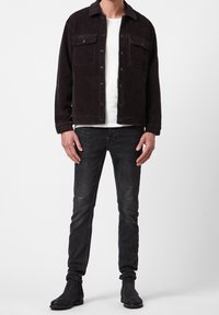 Man som bär en svart cordjacka, vit t-shirt, mörkgrå slim-fit jeans och svarta ankelboots som står mot en enhetlig bakgrund.